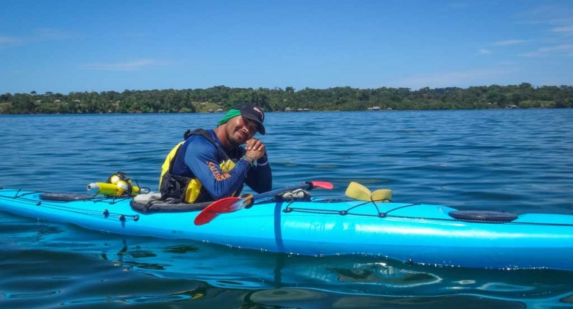 A person takes a break from kayaking on blue water to smile at the camera. In the background, there is a tree-lined shore.